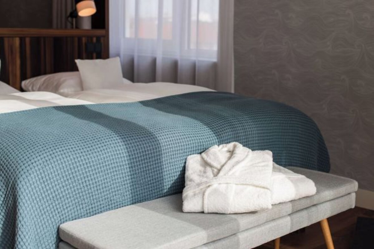 A neatly made bed with a blue and white checkered bedspread, a white pillow, and a folded white towel on a grey bench beside the bed.
