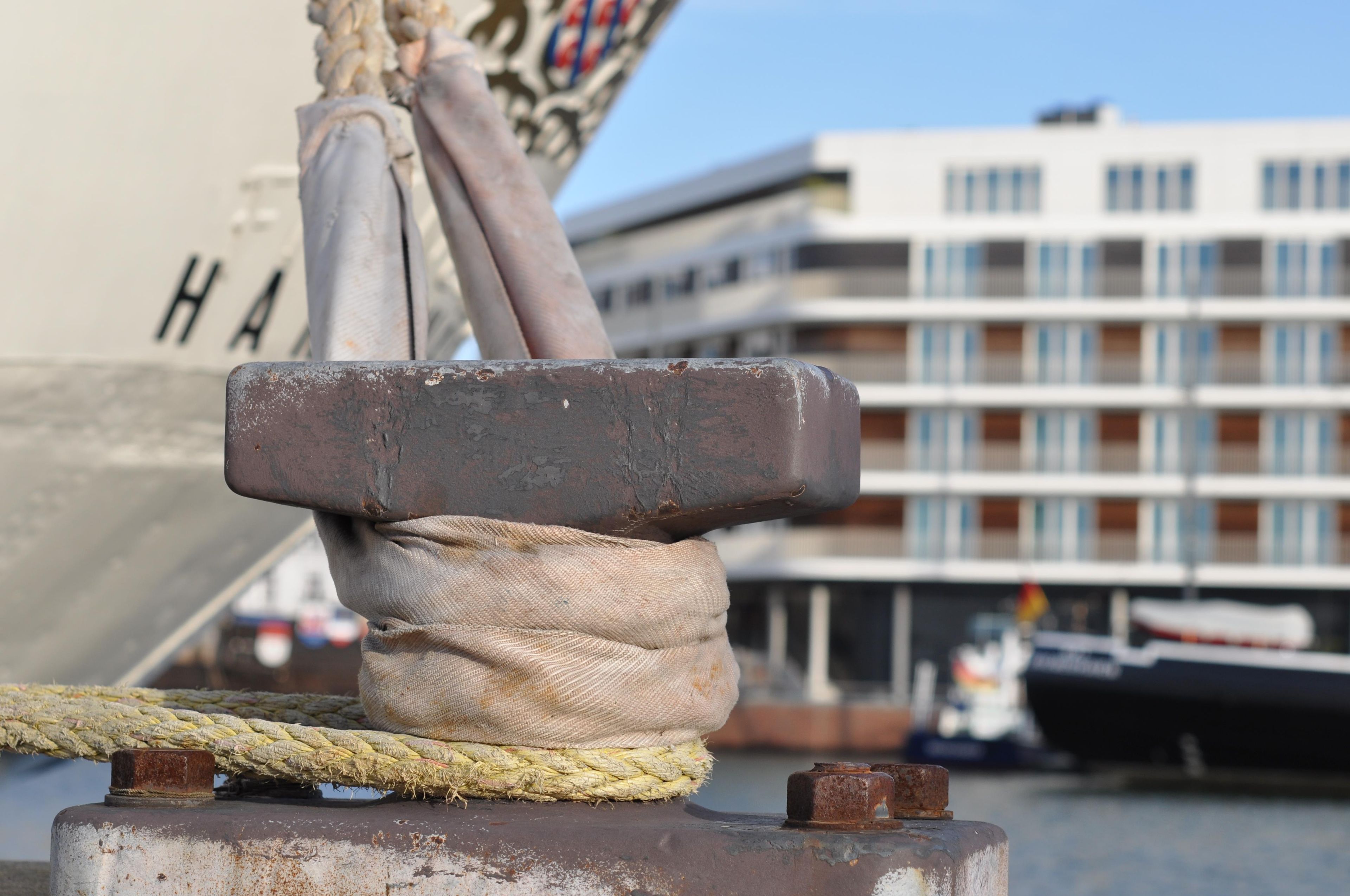 Primer plano de un ancla de barco con una cuerda y un bloque metálico, con un edificio y una embarcación al fondo.