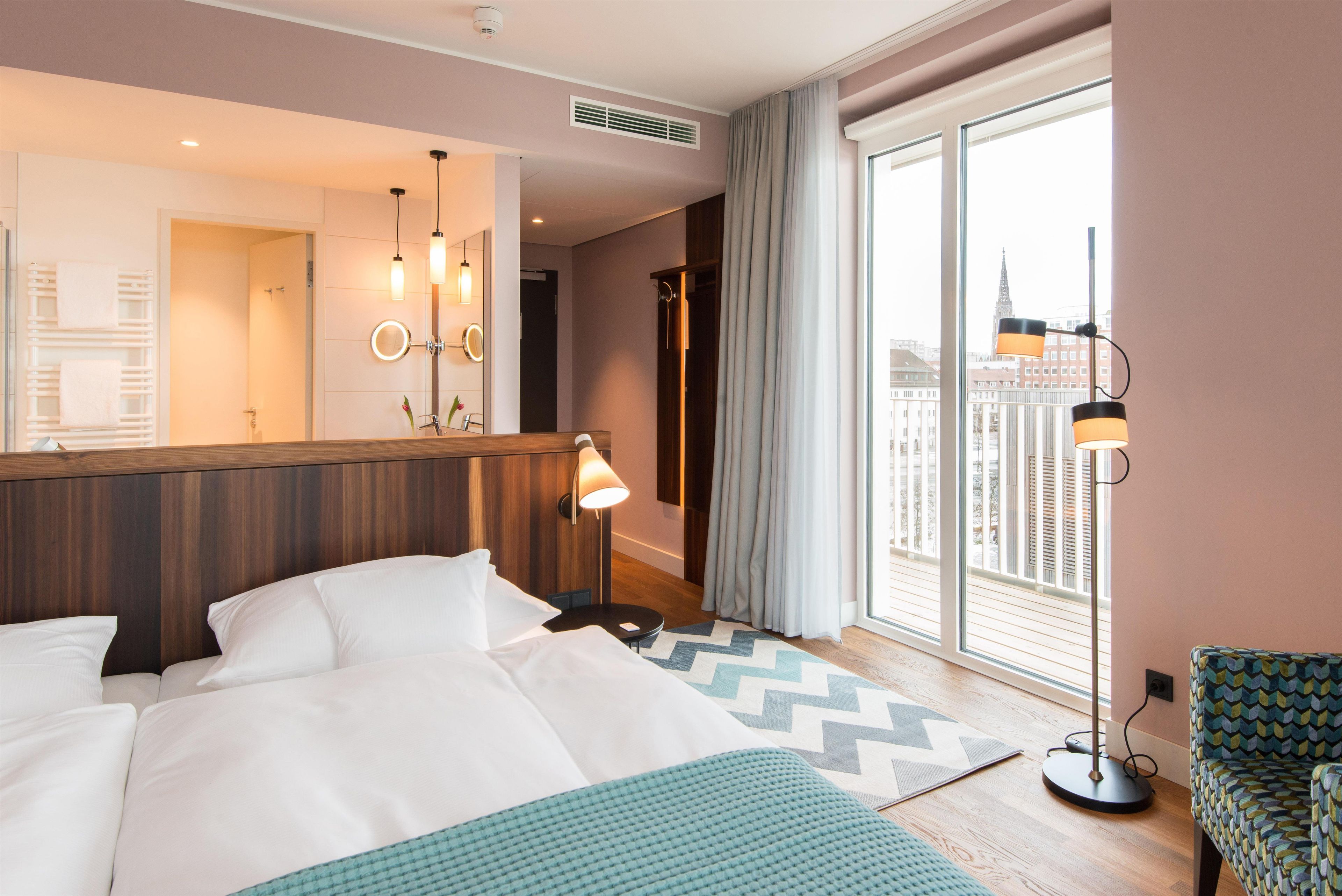 Habitación de hotel con una cama grande, un espejo, una mesita de noche, una lámpara, una ventana con vistas a la ciudad y una alfombra estampada.