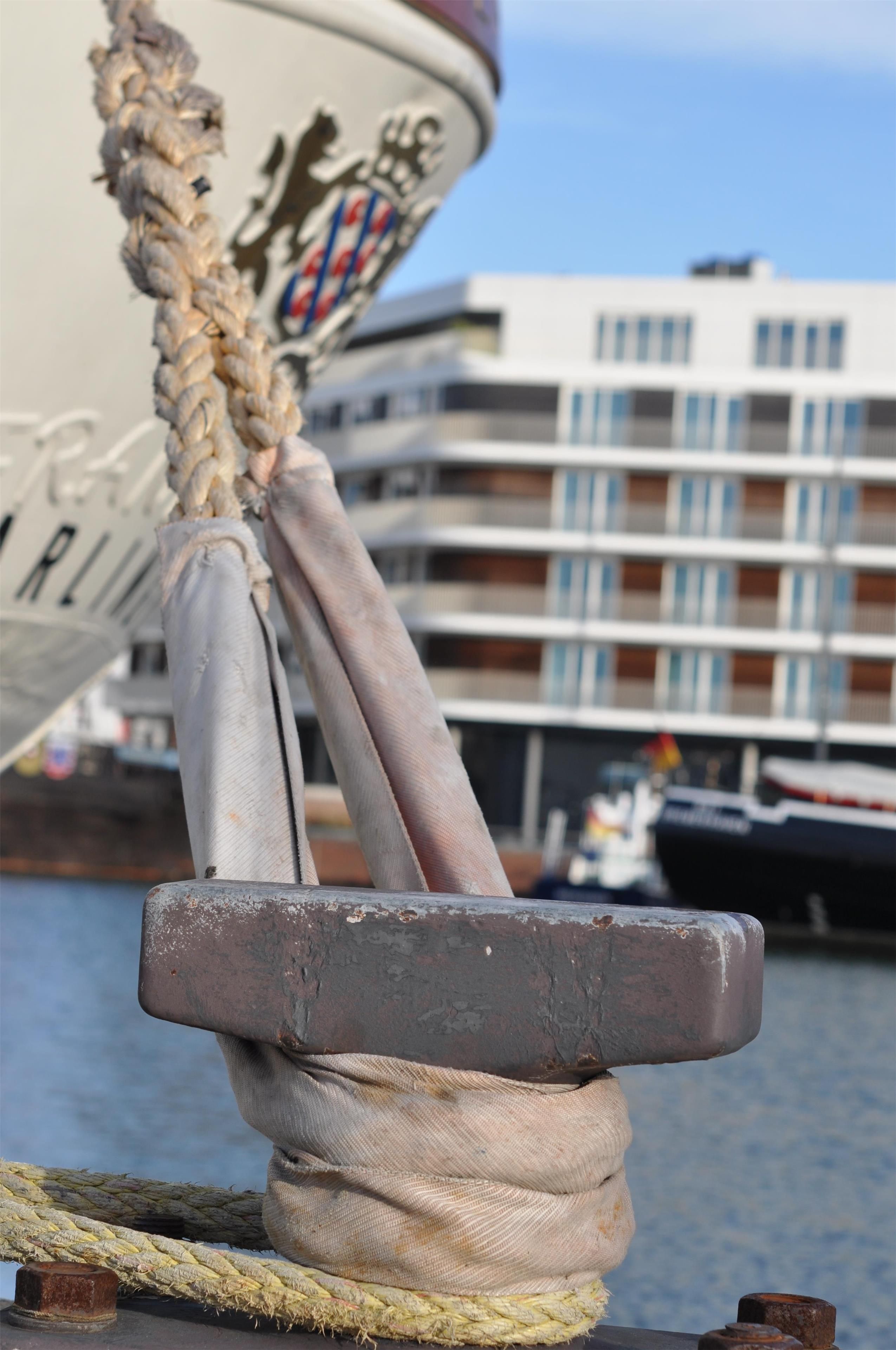 A ship's anchor with a rope and a metal block, a building with multiple windows in the background.