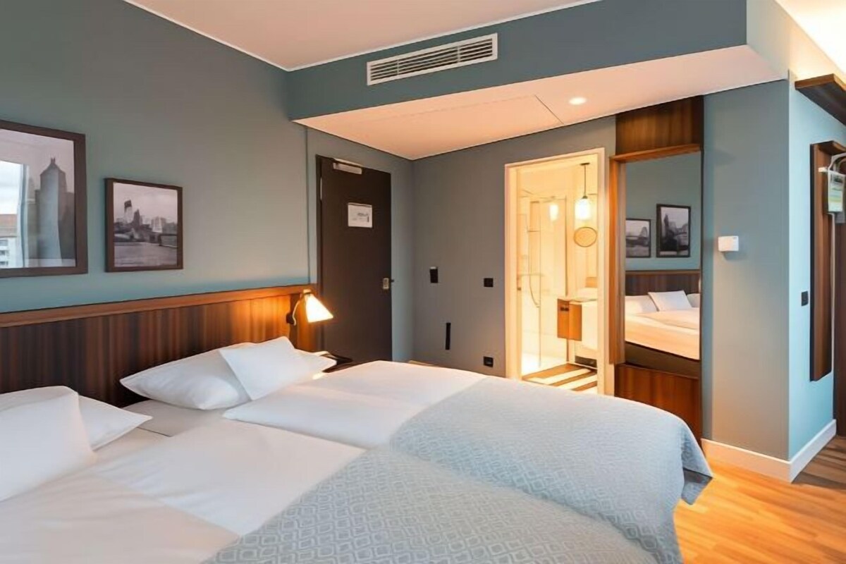 A hotel room with two beds, a wooden headboard, wall-mounted artwork, a door with a sign, and a bathroom visible through an open door.
