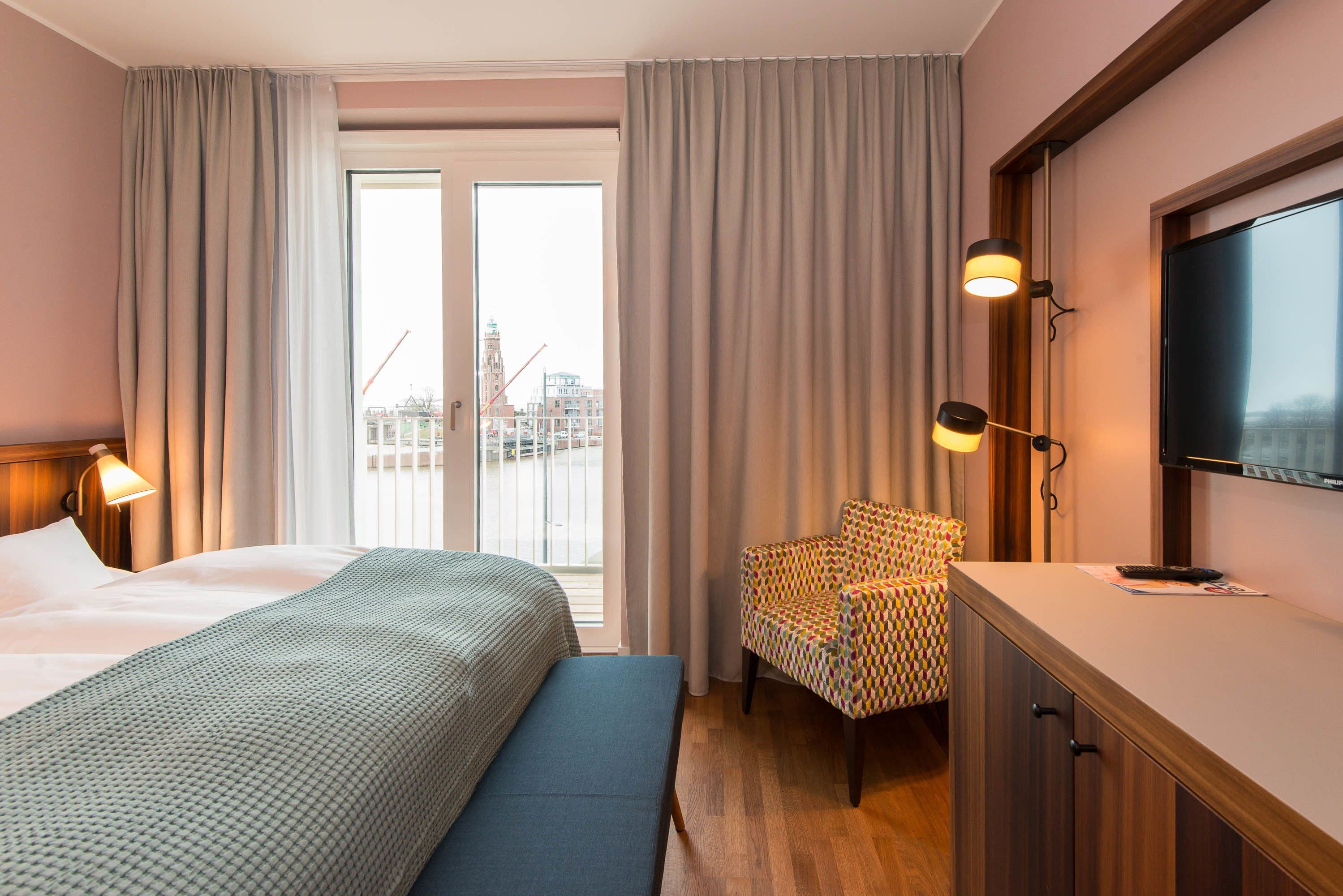 A hotel room with a bed, a bench, a chair, a TV, and a window with curtains.