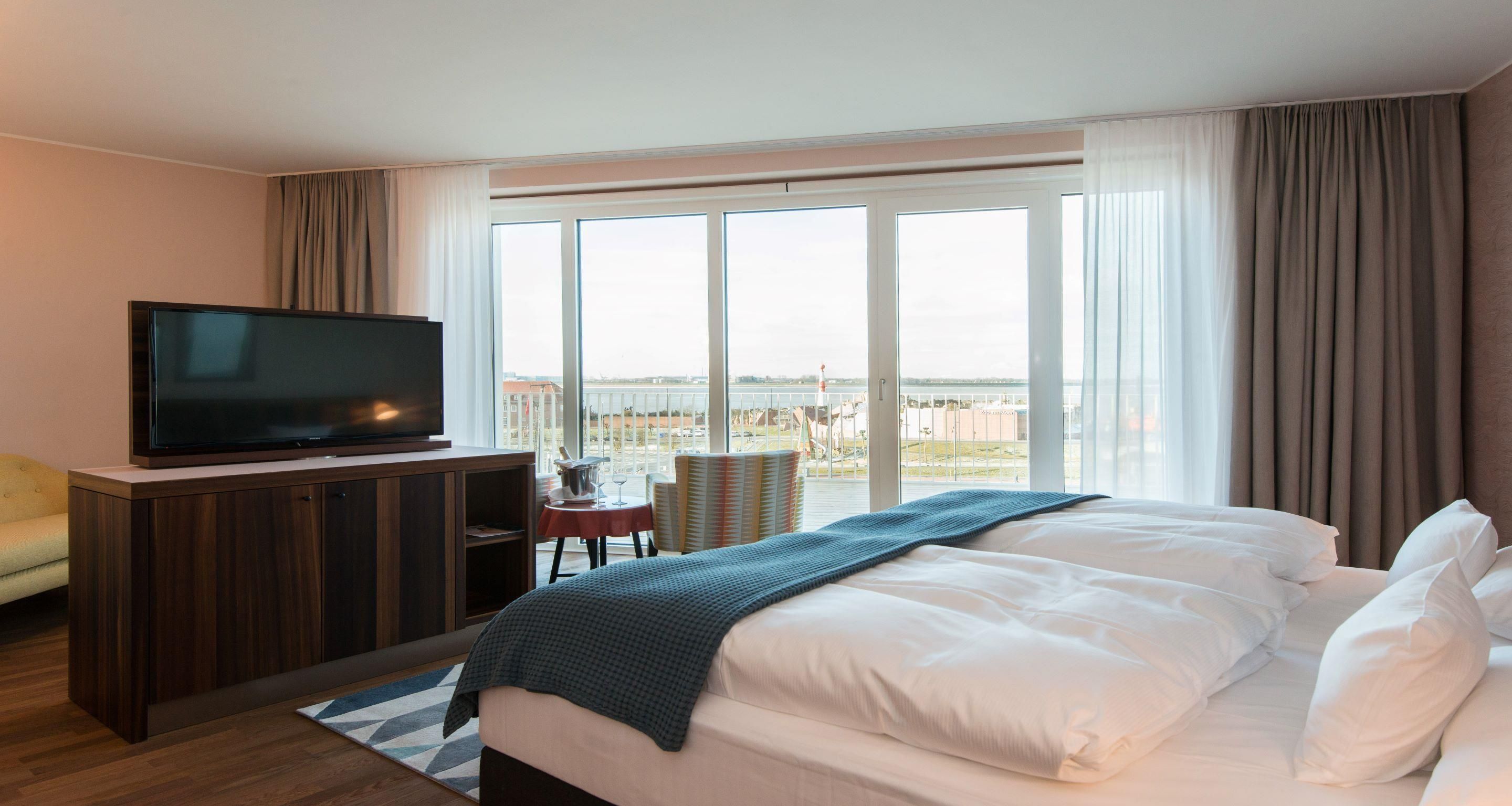Une chambre d’hôtel avec un lit, un téléviseur posé sur un meuble en bois, une petite table avec une chaise, et une vue sur la ville grâce à de grandes fenêtres.