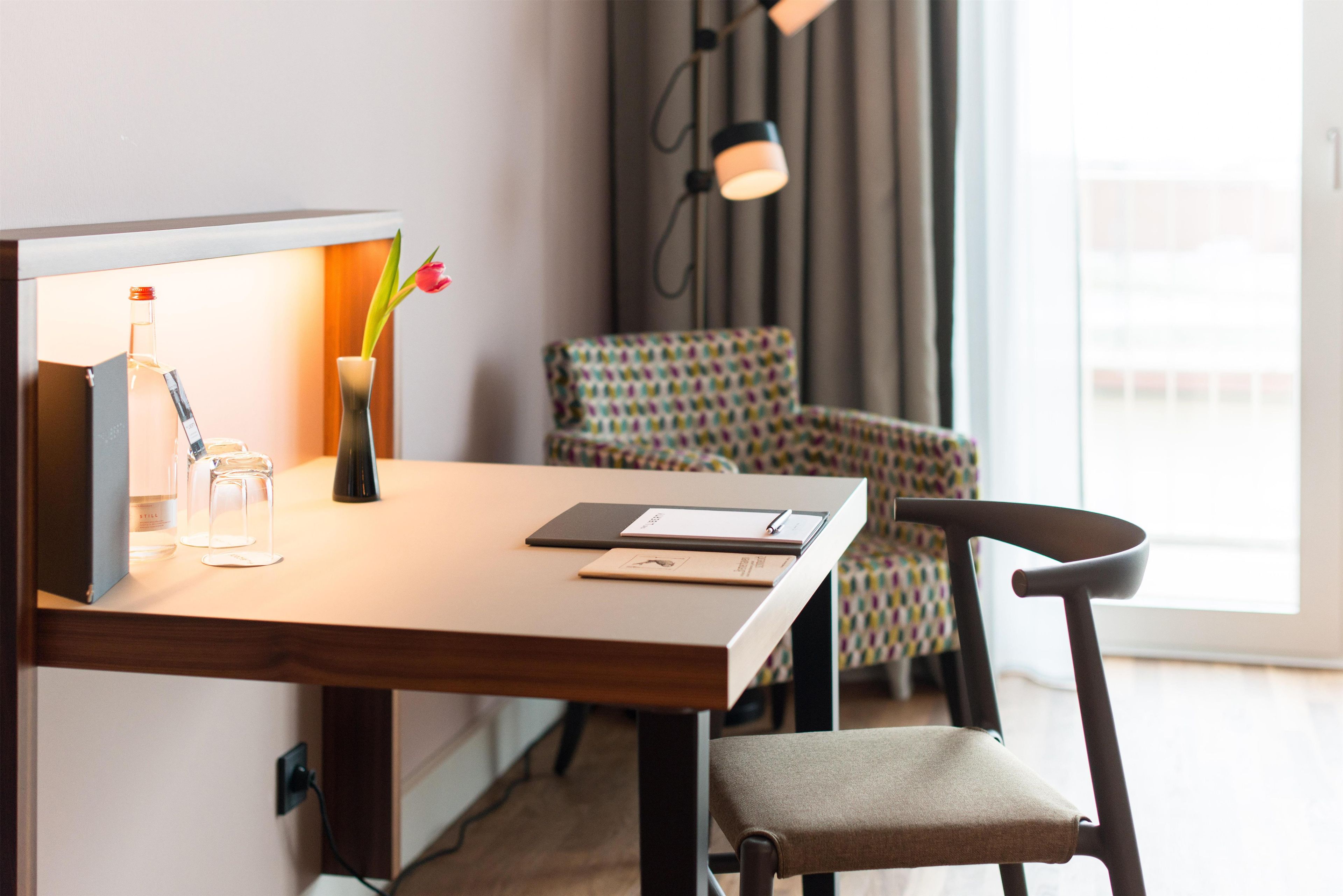 A hotel room with a wooden desk, a chair, a lamp, a vase with a flower, and a window with curtains.