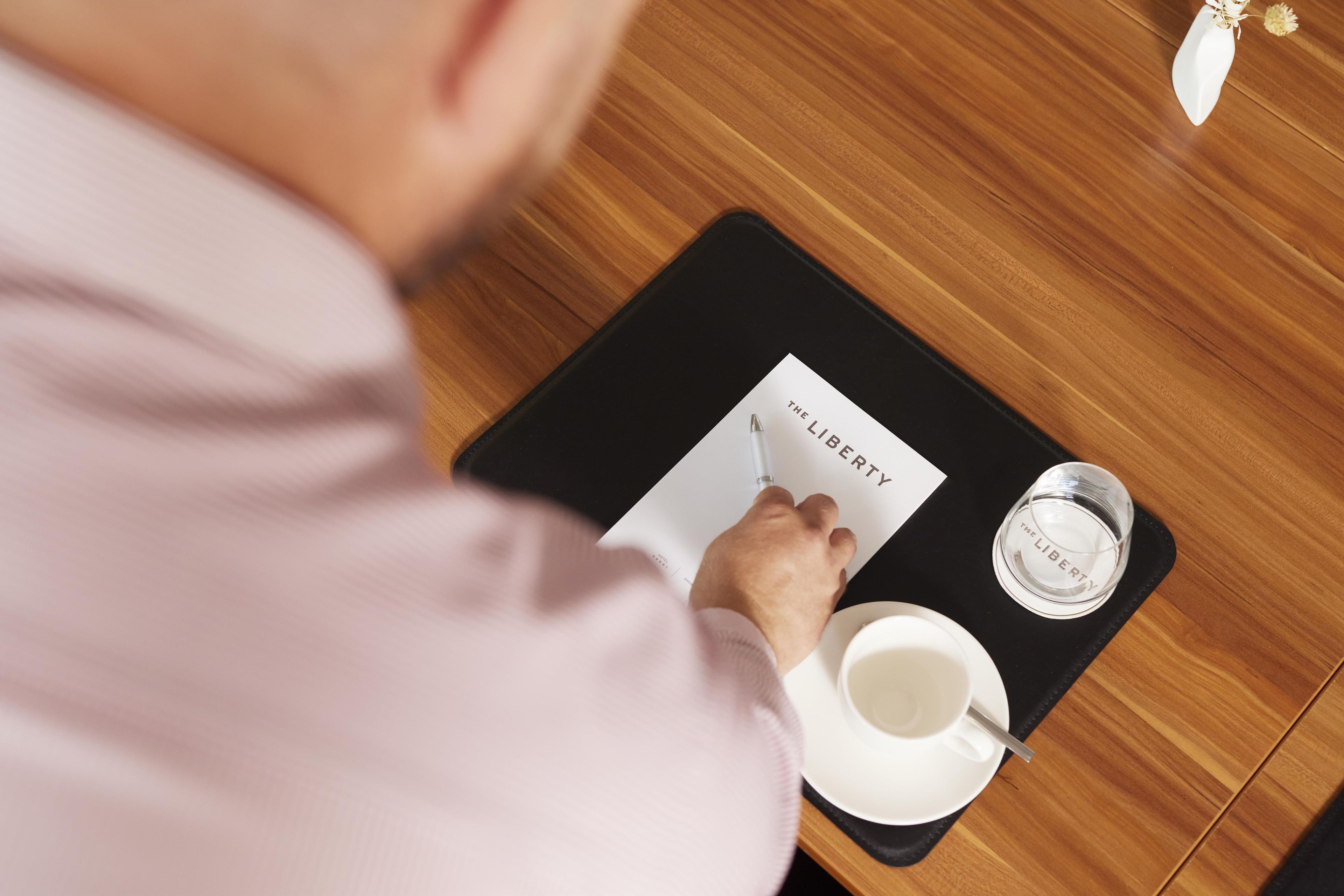 Een persoon schrijft in een notitieboekje met de titel 'The Liberty', met een kopje en een glas op een houten tafel.