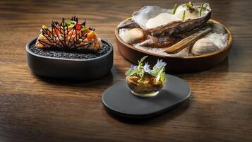 Una mesa de madera con tres platos: un tazón negro con un arreglo decorativo, un bol de madera con mariscos y conchas, y un recipiente pequeño con vegetales verdes y salsa.