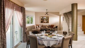 A dining area with a table set for six, a sofa, and a staircase in the background. - Villa Artemis, Wine Dark Sea Villas (Malevizi)