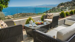 A terrace with wicker furniture, a table with a teapot and cups, and a view of the sea and hills. - Villa Artemis, Wine Dark Sea Villas (Malevizi)