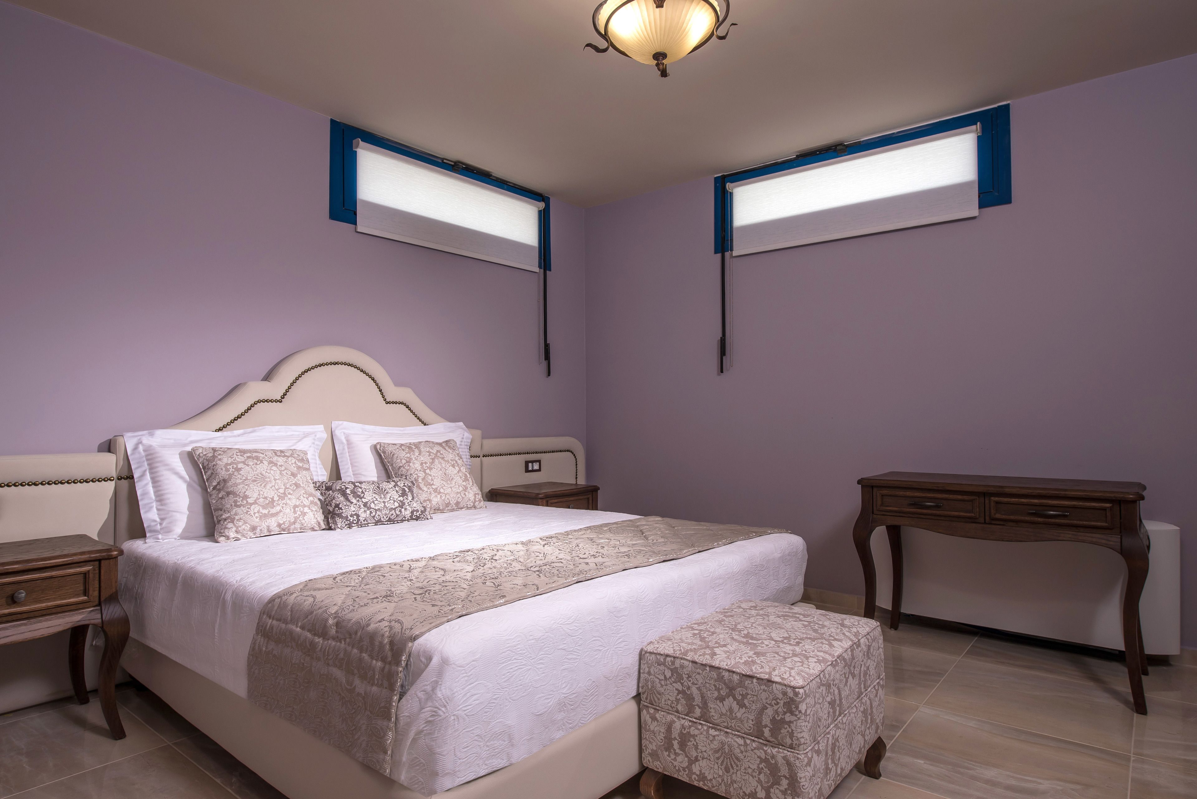 Un dormitorio con una cama grande, mesitas de noche, un tocador y dos ventanas con persianas.