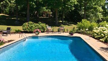 A rectangular swimming pool with a wooden deck, surrounded by lush greenery and a pergola.