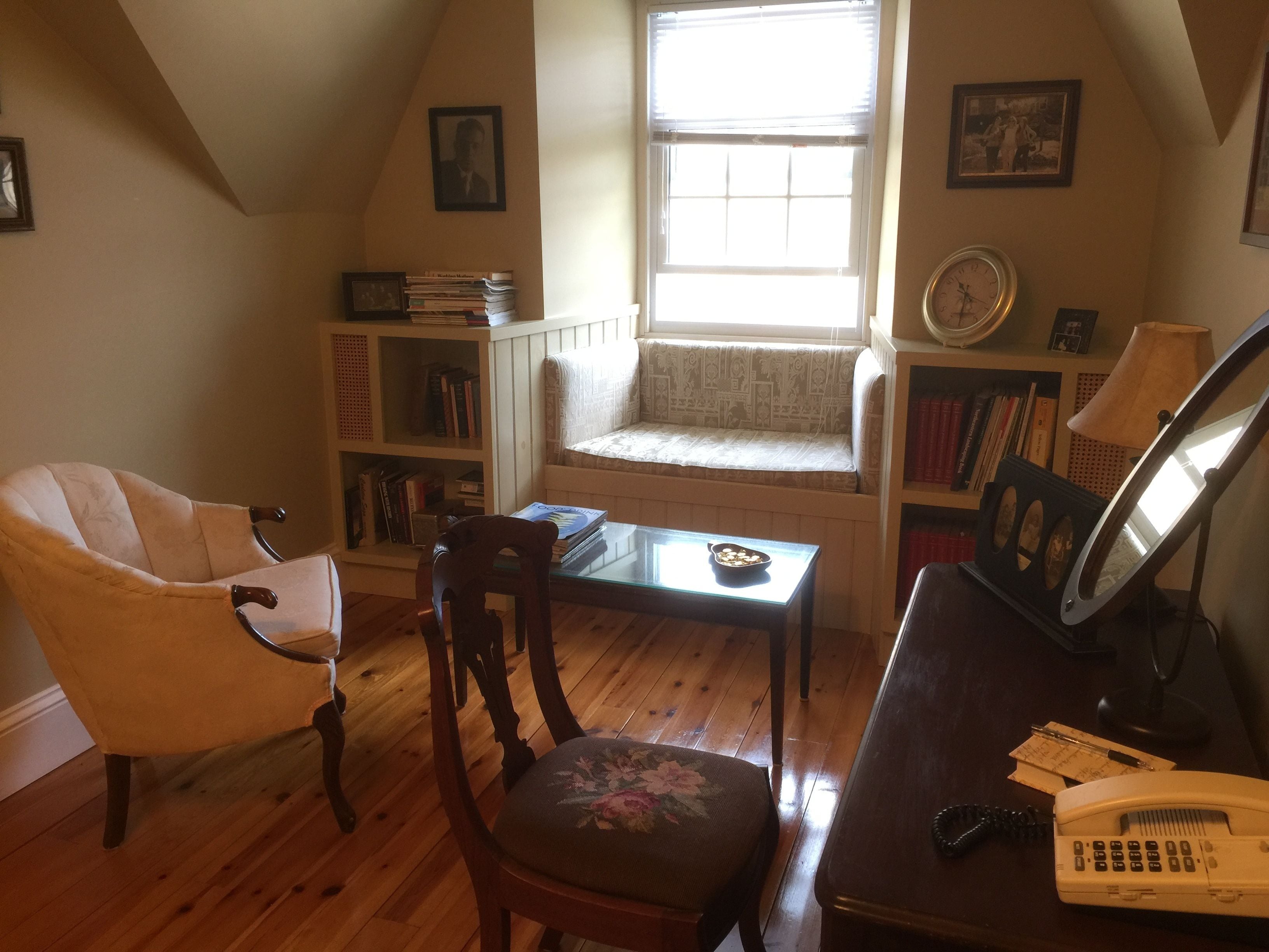 A cozy room with a wooden floor, a window seat, a bookshelf, a desk with a telephone, and a chair.