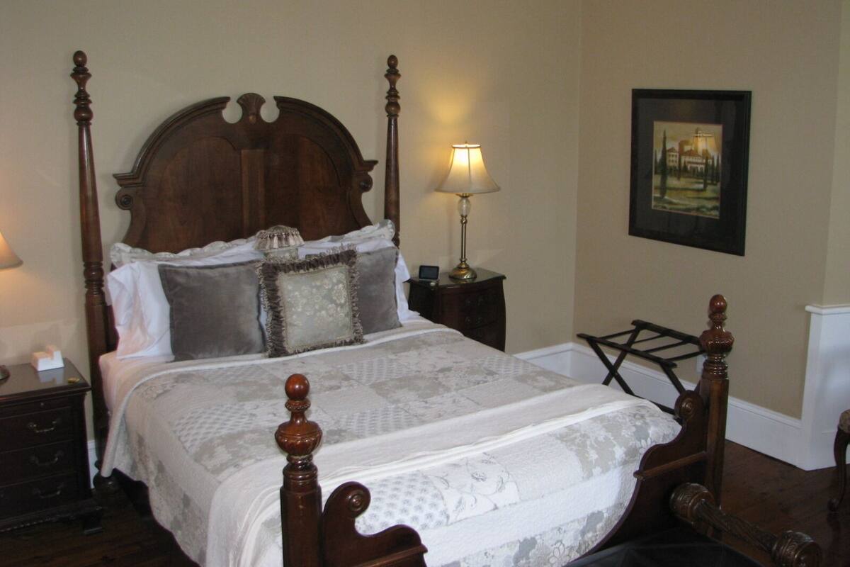 A four-poster bed with a wooden headboard, a bedside table with a lamp, and a framed picture on the wall.
