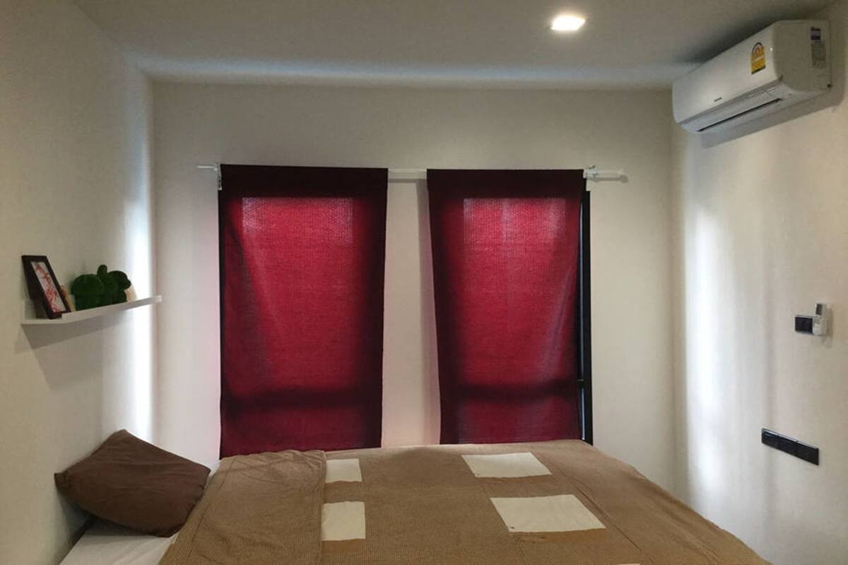 A bedroom with a bed, red curtains, a brown pillow, a white shelf with a plant, and an air conditioning unit.