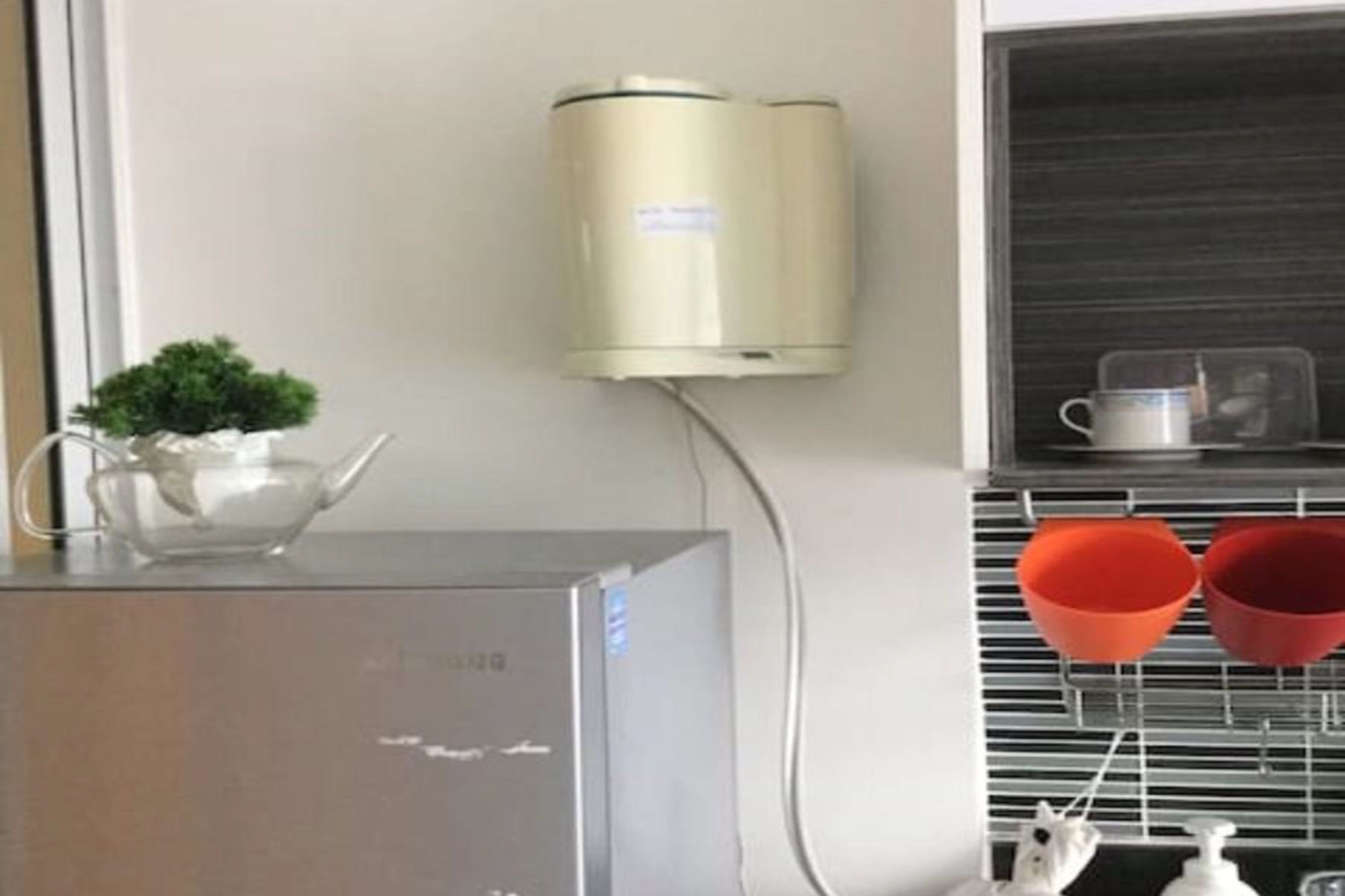 A kitchen with a stainless steel refrigerator, a wall-mounted coffee maker, and a shelf with kitchen utensils and a small potted plant.