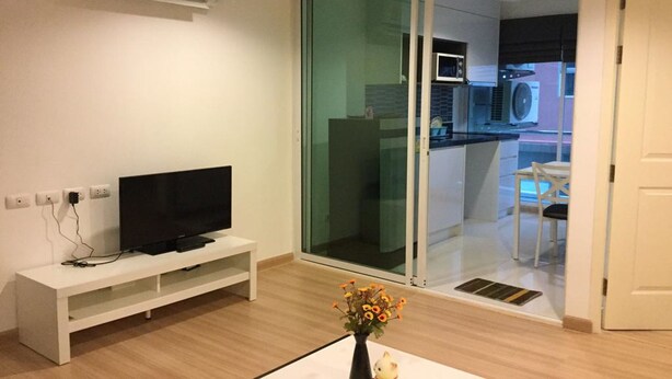 A modern living room with a flat-screen TV on a white stand, a vase with flowers, and a sliding glass door leading to a kitchen area. - Chateau In Town By Renvio (Bangkok)