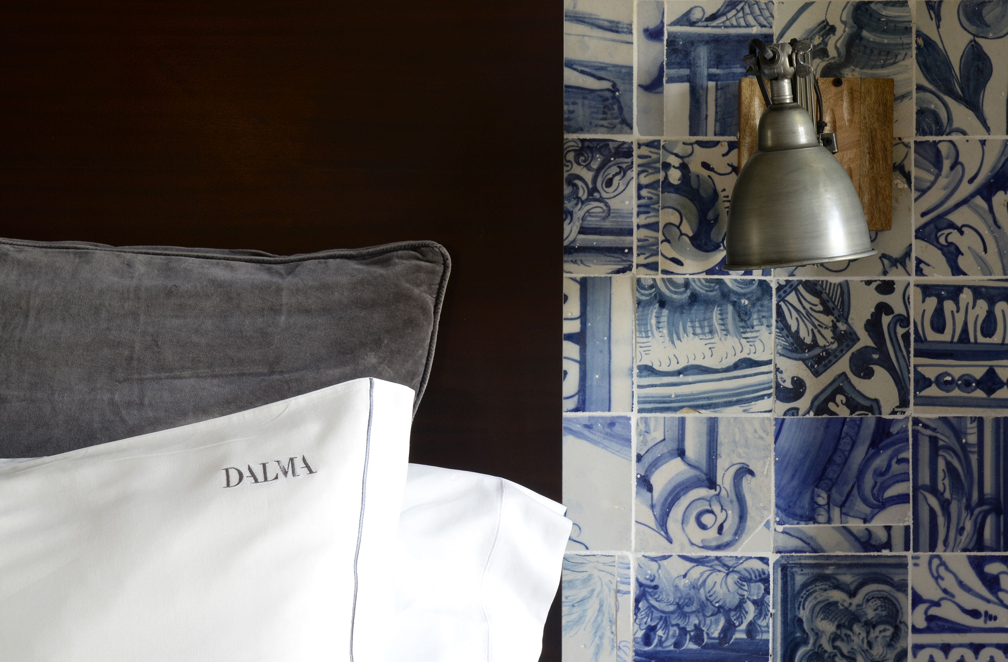 A bed with a pillow labeled "DALMA" and a metal lamp on a tiled wall with blue and white patterns.