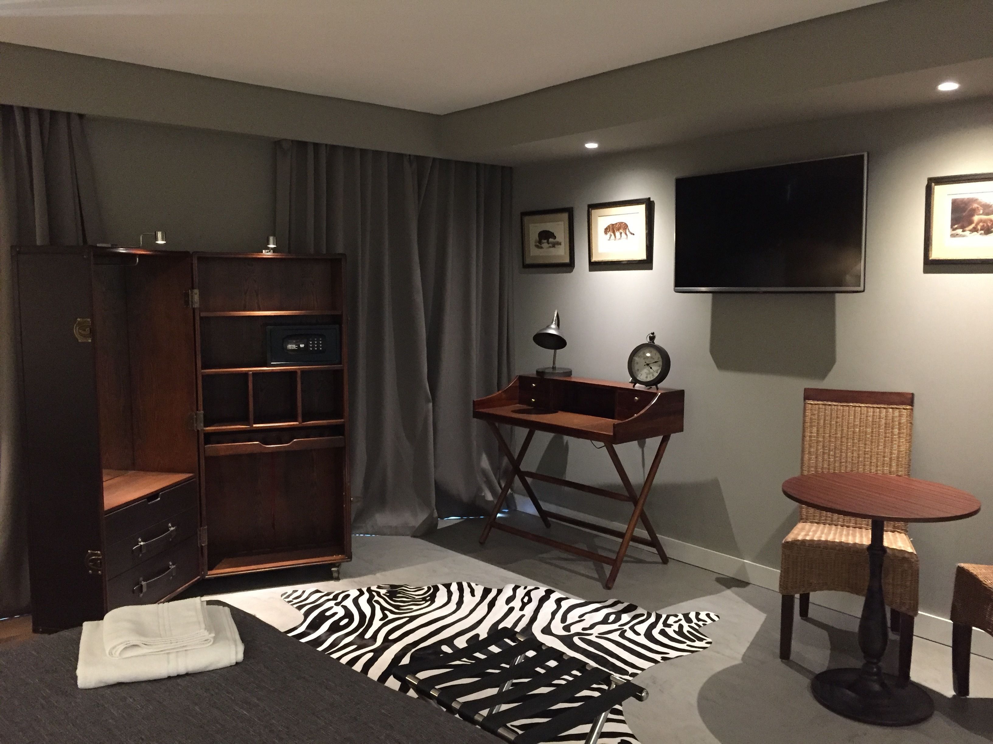 Une chambre d’hôtel avec une armoire en bois, un tapis à motifs zébrés, une petite table ronde avec une chaise, un bureau avec une lampe et un téléviseur à écran plat fixé au mur.