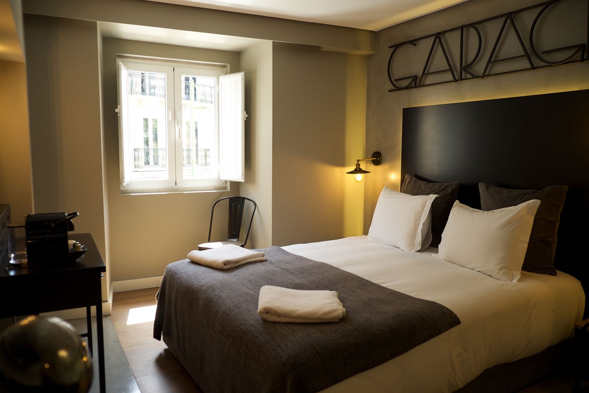 A hotel room with a bed, a desk, a chair, a window, and a wall-mounted light.