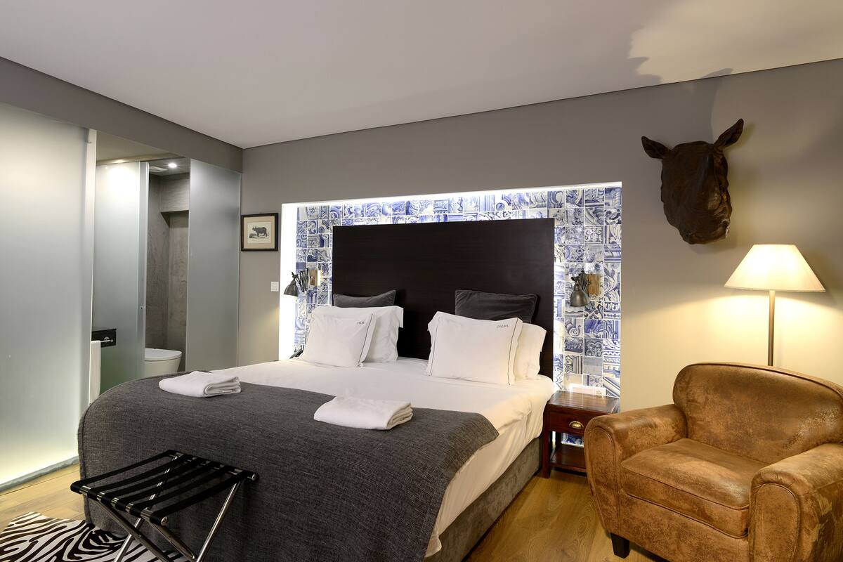 Une chambre d’hôtel moderne avec un grand lit, une tête de lit à motifs bleus et blancs, une décoration murale représentant une tête d’animal, un fauteuil en cuir et une table de chevet avec une lampe.