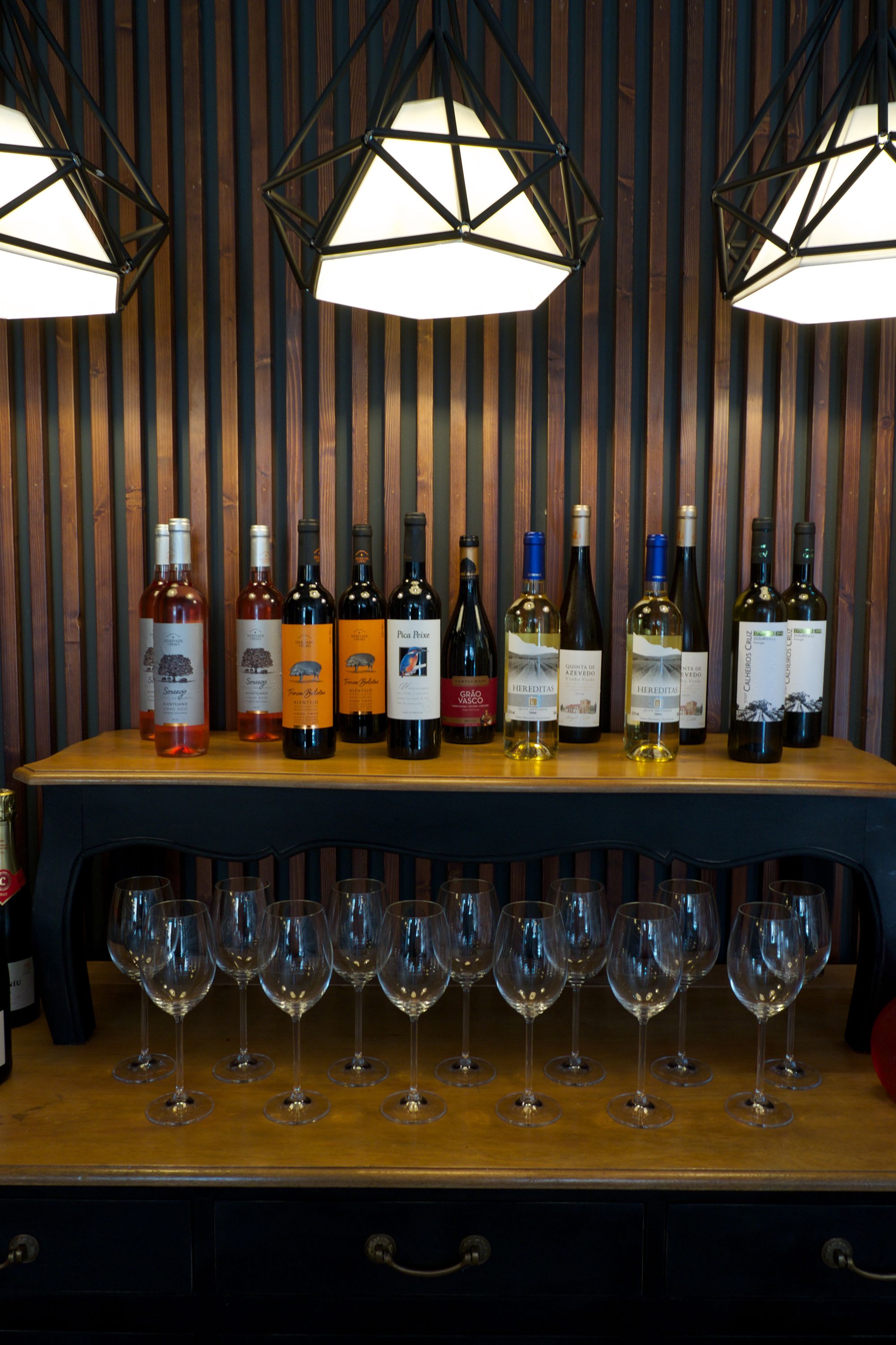 Disposição para degustação de vinhos, com garrafas e copos sobre mesa de madeira, em frente a uma parede listrada.