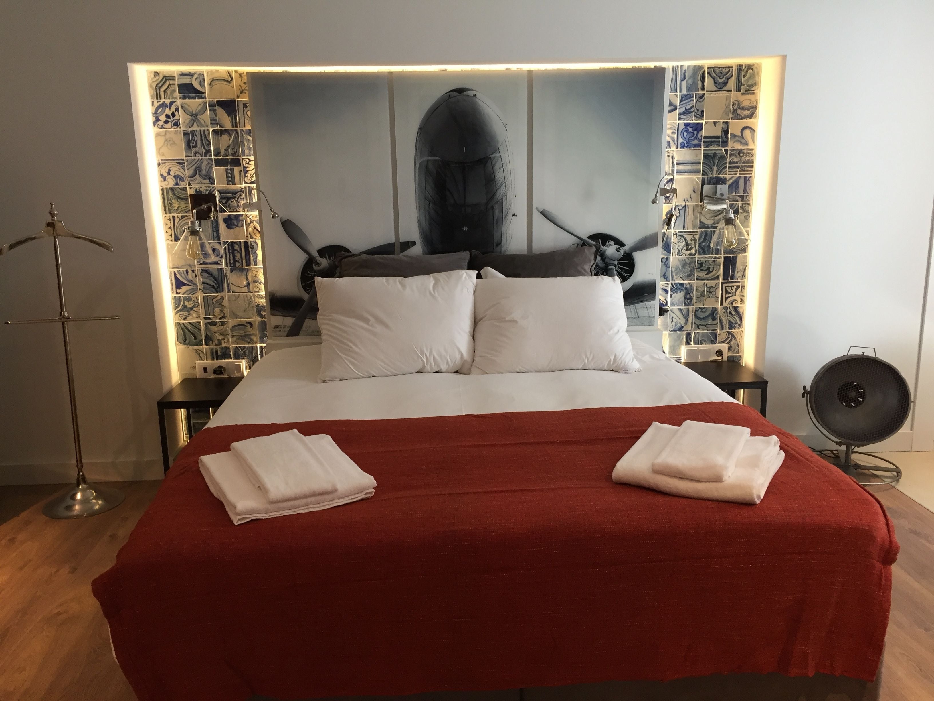 A neatly made bed with white linens and a red blanket, a large framed artwork above the bed, and a small table with a lamp beside it.