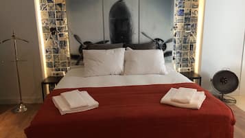 A neatly made bed with white linens and a red blanket, a large framed artwork above the bed, and a small table with a lamp beside it.