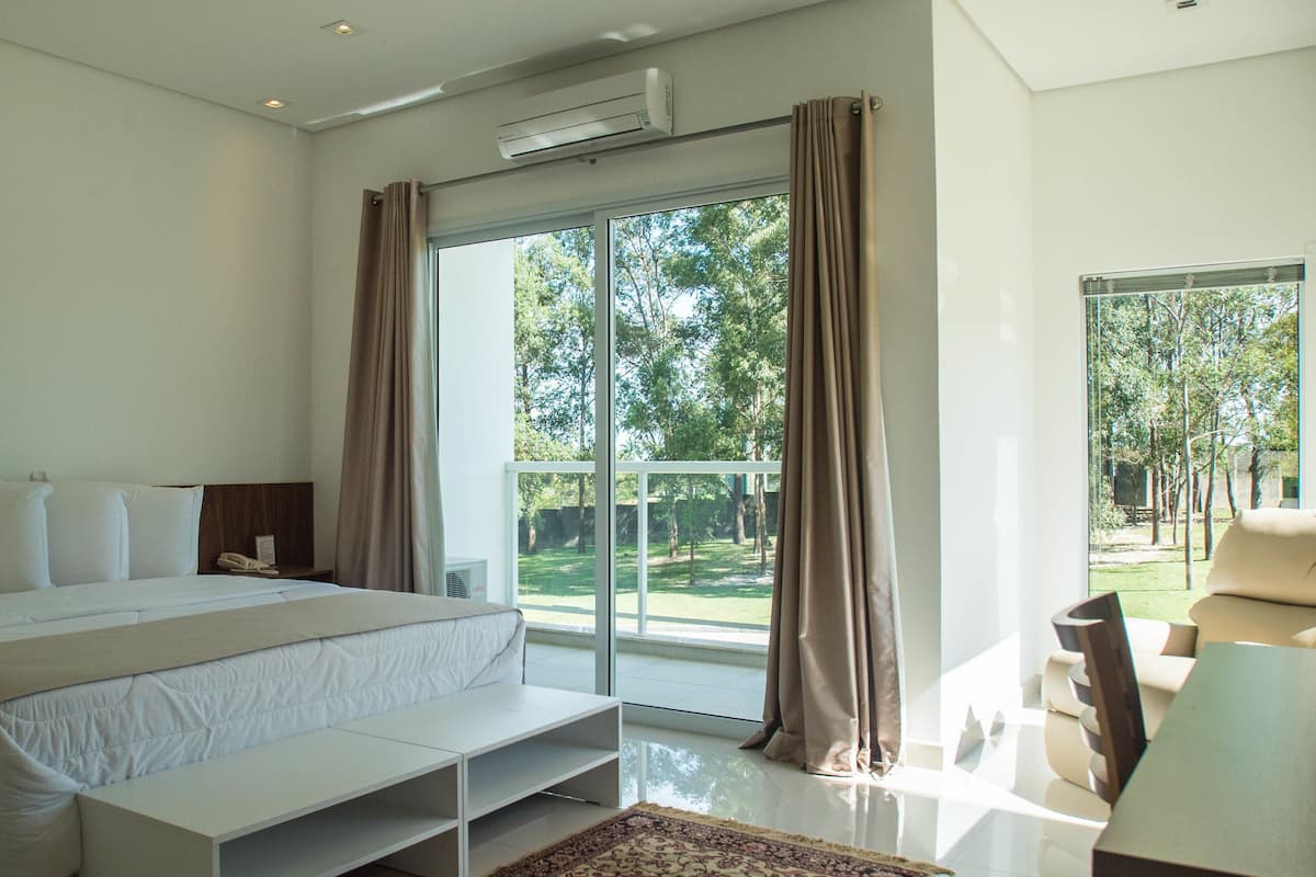 A modern bedroom with a large bed, a bedside table, an air conditioning unit, and a view of a garden through a sliding glass door.