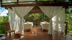 A wooden deck with four tables, each with a white tablecloth and a candle. There is a wicker chair and a wooden bucket on the deck.