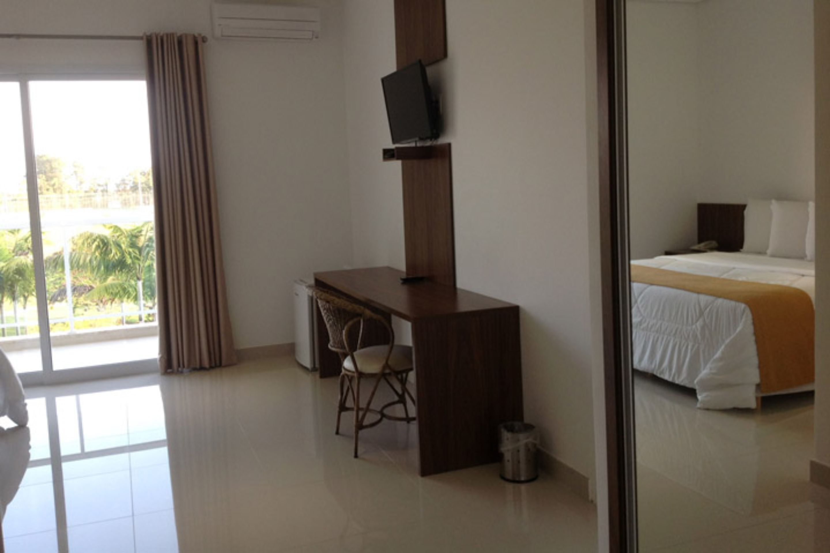 A hotel room with a bed, a desk, a chair, a television, and a view of palm trees through the window.