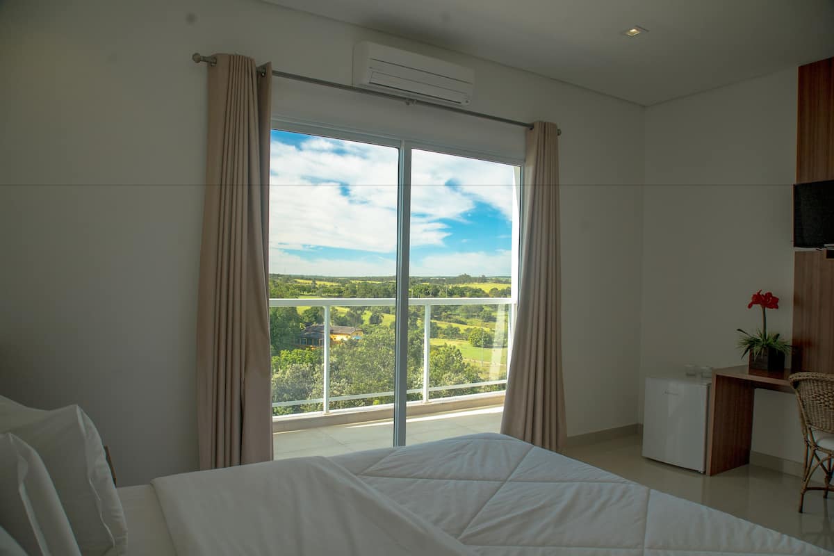 A bedroom with a large bed, a desk, and a view of a green landscape through a sliding glass door.