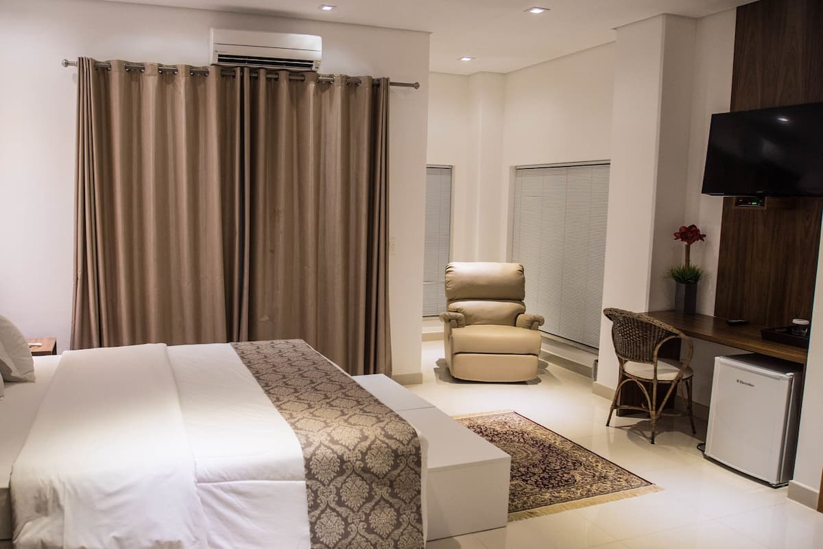 A hotel room with a large bed, a desk with a television, a chair, and an air conditioning unit.