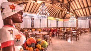 Uma sala de jantar ampla, com várias mesas e cadeiras, teto de madeira alto e lustres.