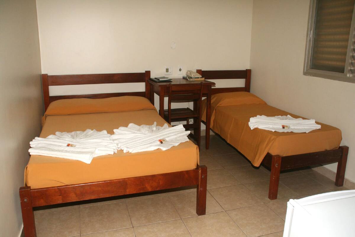 A room with two wooden beds, each with an orange bedspread and white towels. There is a small wooden nightstand with a lamp and a book.