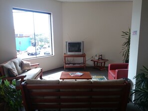 Un salon avec un canapé, un fauteuil rouge, une table basse, un téléviseur posé sur un meuble et une fenêtre donnant sur la rue.