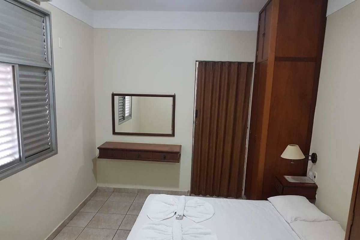 A hotel room with a bed, a bedside table, a mirror, a window with blinds, a wooden bench, and a curtain.