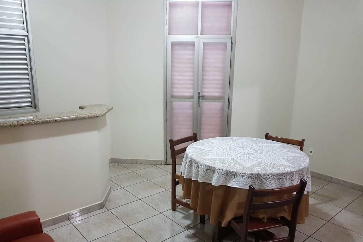 A small, tiled room with a round table covered by a white lace tablecloth, wooden chairs, a window with blinds, and a door with frosted glass panels.