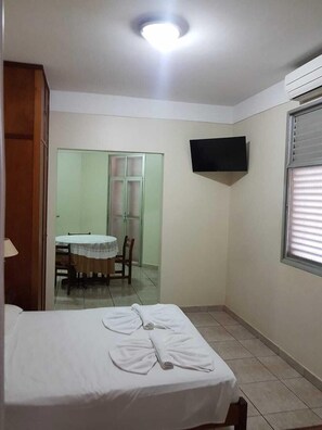 Quarto de hotel com cama, mesa redonda com cadeiras, televisão presa na parede e janela com persianas.