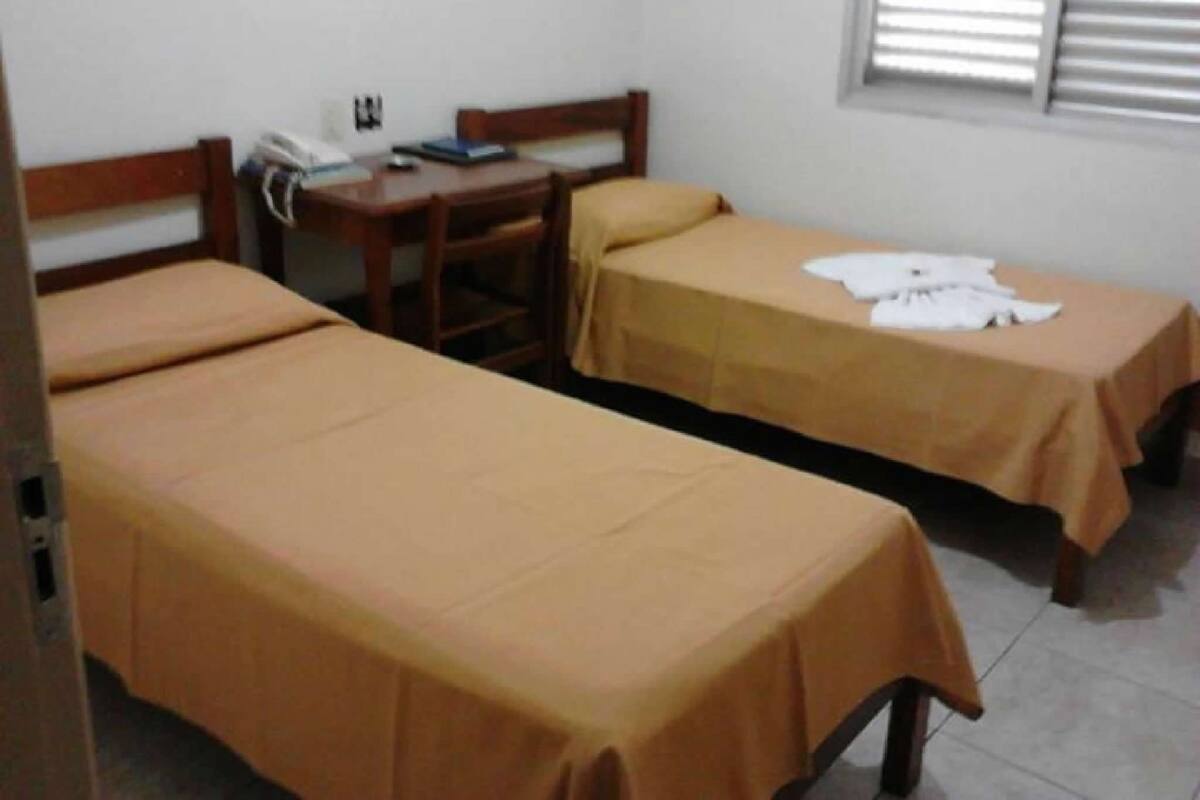 A room with two single beds, a wooden desk, a chair, and a window with blinds.