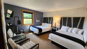 A hotel room with two beds, a desk, a chair, a TV, and a window with a view of greenery.