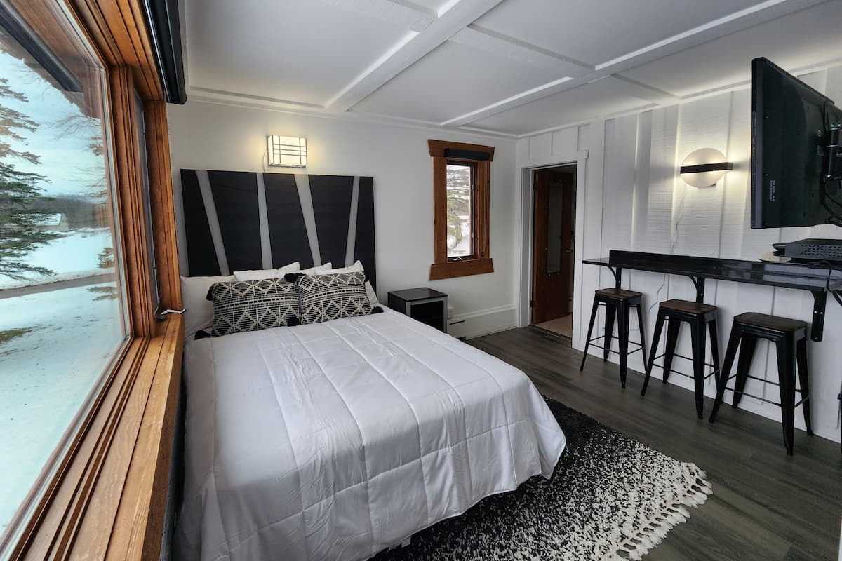 Un dormitorio con una cama grande, un escritorio con taburetes, un televisor y una ventana con vistas a árboles.