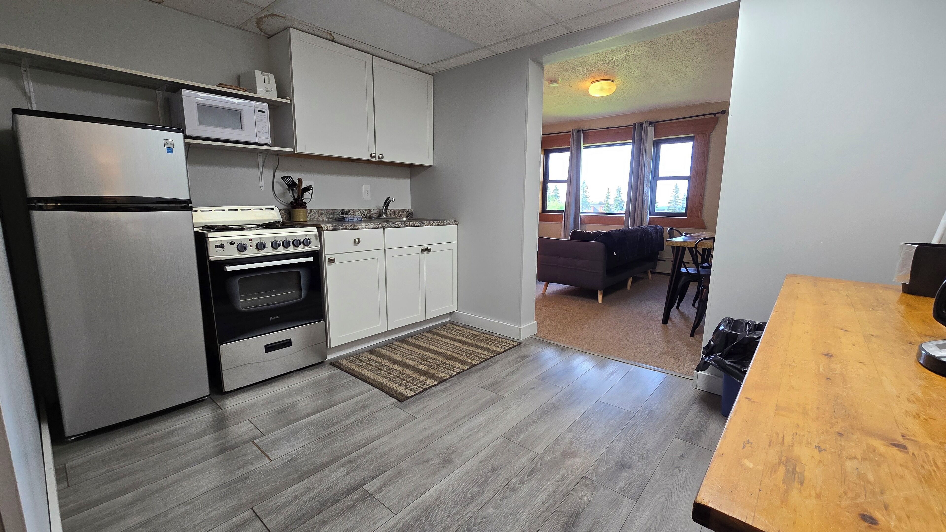 A kitchen with a refrigerator, stove, microwave, and cabinets. There is a wooden table and a rug in the dining area.