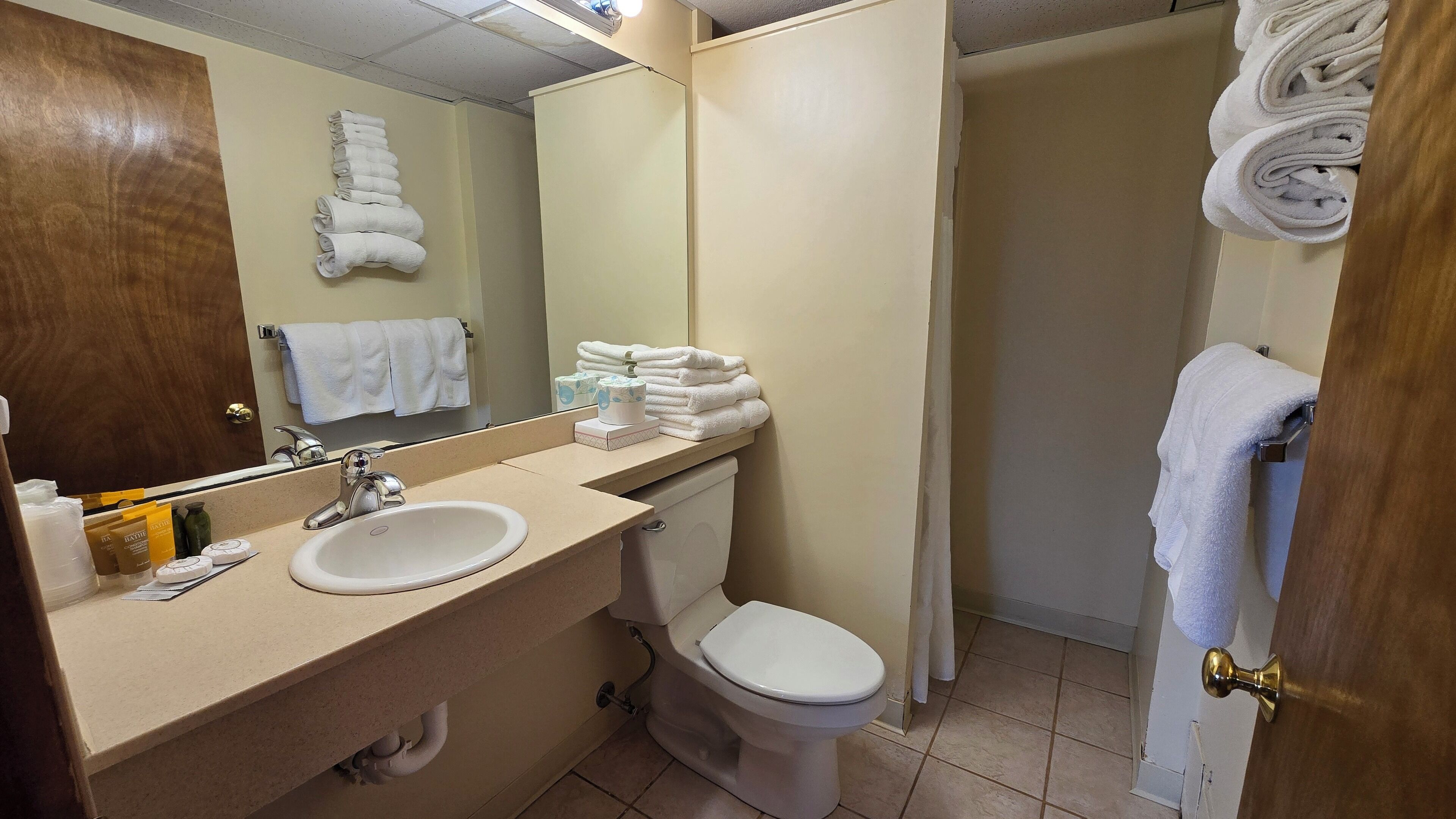 A bathroom with a sink, toilet, and a mirror. There are towels stacked on the wall and a door with a towel hanging on it.