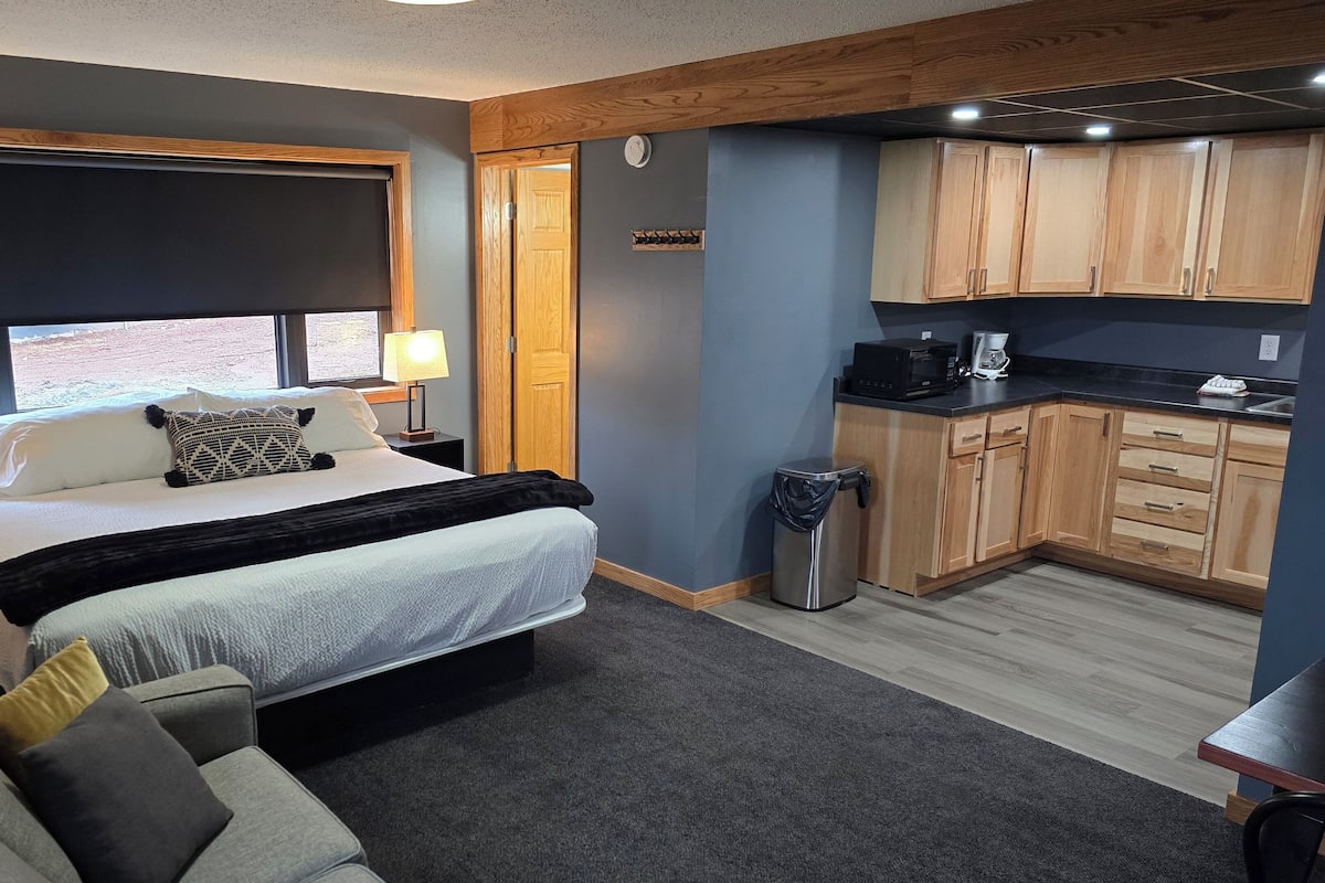 Un dormitorio con cama, lámparas de noche, una ventana con vistas, una pequeña kitchenette con armarios de madera y un cubo de basura.