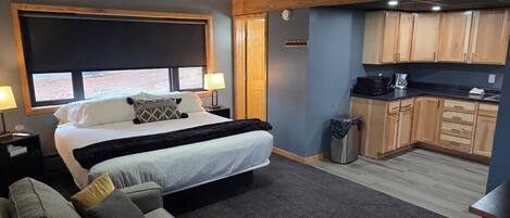 A bedroom with a bed, bedside lamps, a window with a view, a small kitchenette with wooden cabinets, and a trash can.