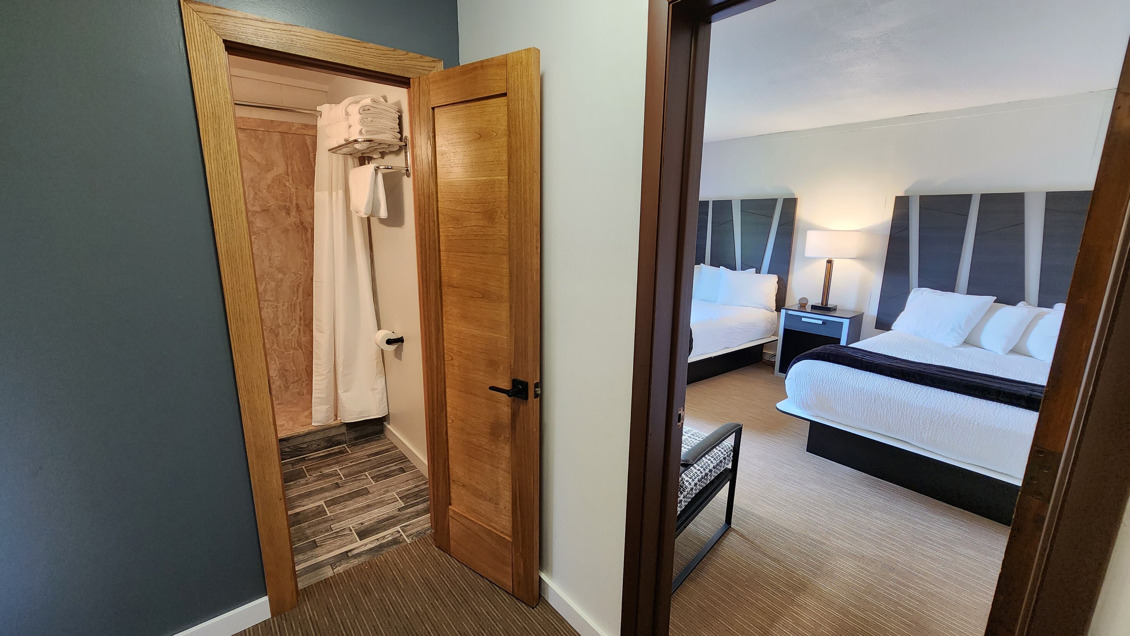 Habitación de hotel con puerta de madera, zona de ducha, dos camas con ropa blanca, mesita de noche con lámpara y un pequeño banco.