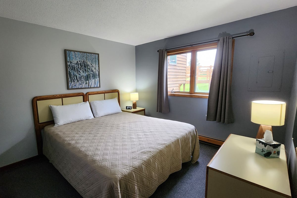Habitación de hotel ordenada con una cama grande, una mesita de noche con lámpara, una ventana con cortinas y un cuadro en la pared.
