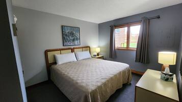 A neatly arranged hotel room with a large bed, a bedside table with a lamp, a window with curtains, and a painting on the wall.