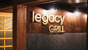 A sign for 'Legacy Grill' with a modern design, featuring a dark textured wall and a wooden panel with a geometric pattern.
