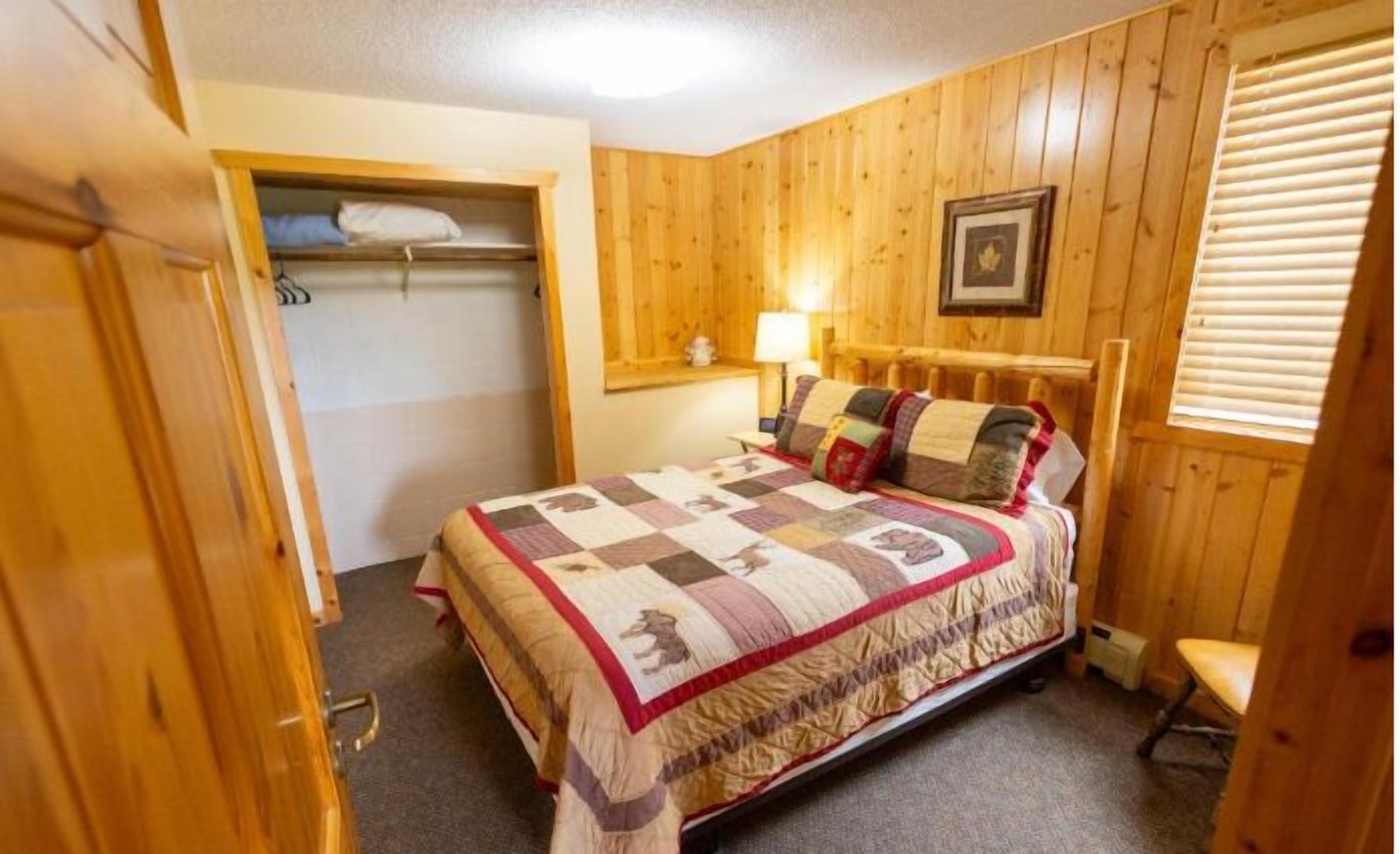 A wooden-paneled bedroom with a bed, a nightstand, a lamp, and a window with blinds.