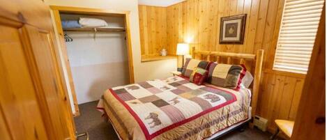 A wooden-paneled bedroom with a bed, a nightstand, a lamp, and a window with blinds.